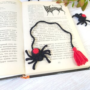 K&ouml;nnte beinhalten: Ein schwarzer und roter H&auml;kel-Spinnen-Lesezeichen mit rotem Quasten, das auf einem offenen Buch liegt. Zus&auml;tzliche H&auml;kelspinnen sind um das Buch herum verstreut, was ein handgemachtes Thema schafft.