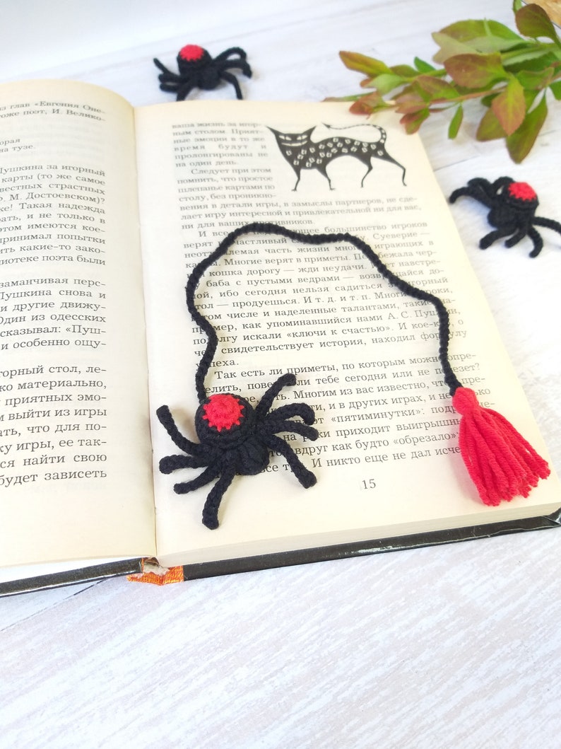 K&ouml;nnte beinhalten: Ein schwarzer und roter H&auml;kel-Spinnen-Lesezeichen mit rotem Quasten, das auf einem offenen Buch liegt. Zus&auml;tzliche H&auml;kelspinnen sind um das Buch herum verstreut, was ein handgemachtes Thema schafft.