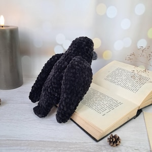 May include: A black crocheted bird figurine with a soft, plush texture, featuring a rounded body and folded wings, positioned next to an open book with visible printed text and a small pinecone on a light-coloured surface near a grey candle.