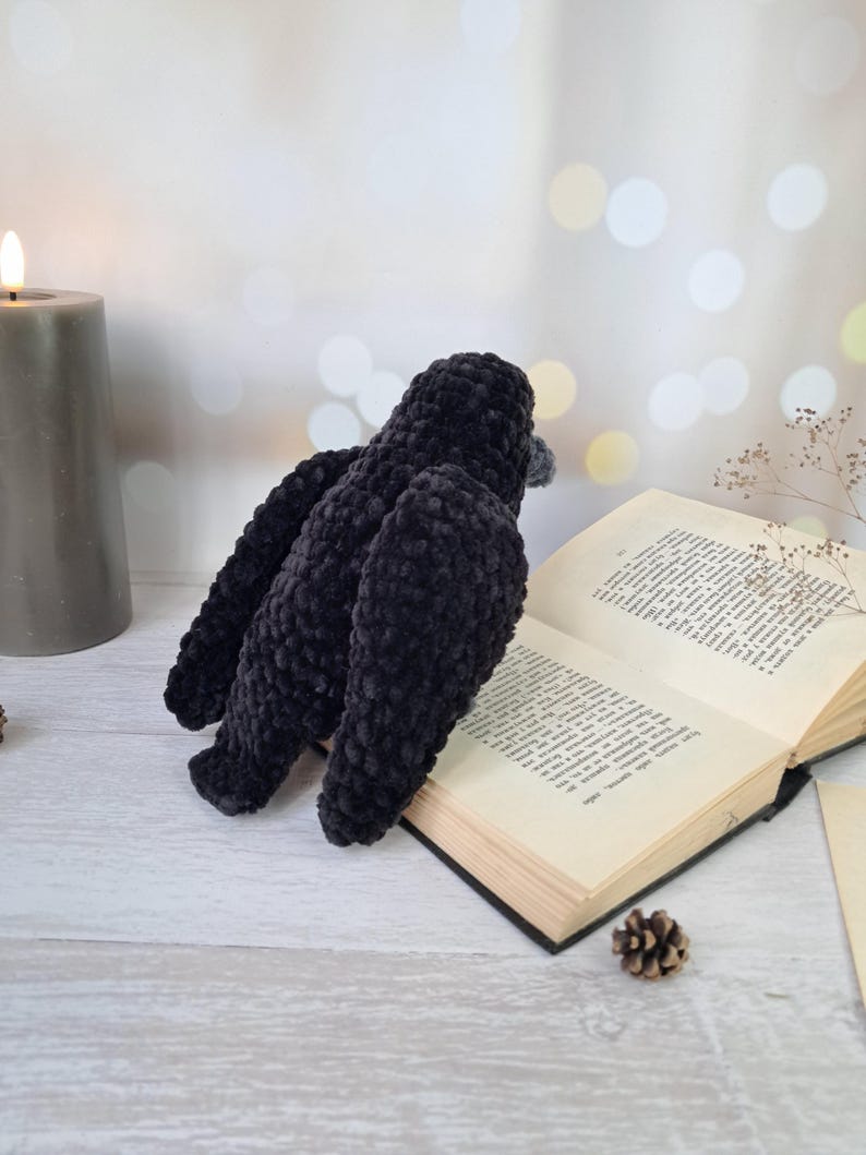 May include: A black crocheted bird figurine with a soft, plush texture, featuring a rounded body and folded wings, positioned next to an open book with visible printed text and a small pinecone on a light-coloured surface near a grey candle.