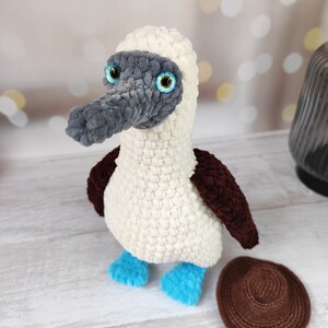 May include: A crocheted blue-footed booby bird plush toy with a gray head, white body, brown wings, and blue feet. The toy is sitting on a white wooden surface with a brown crocheted circle next to it.