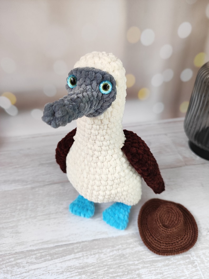 May include: A crocheted blue-footed booby bird plush toy with a gray head, white body, brown wings, and blue feet. The toy is sitting on a white wooden surface with a brown crocheted circle next to it.