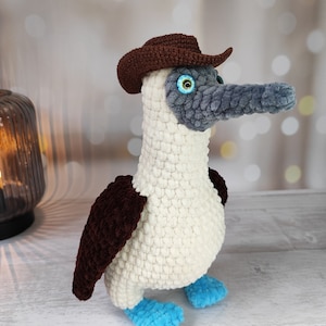 May include: A crocheted blue-footed booby wearing a brown cowboy hat. The bird is white with brown wings and blue feet.