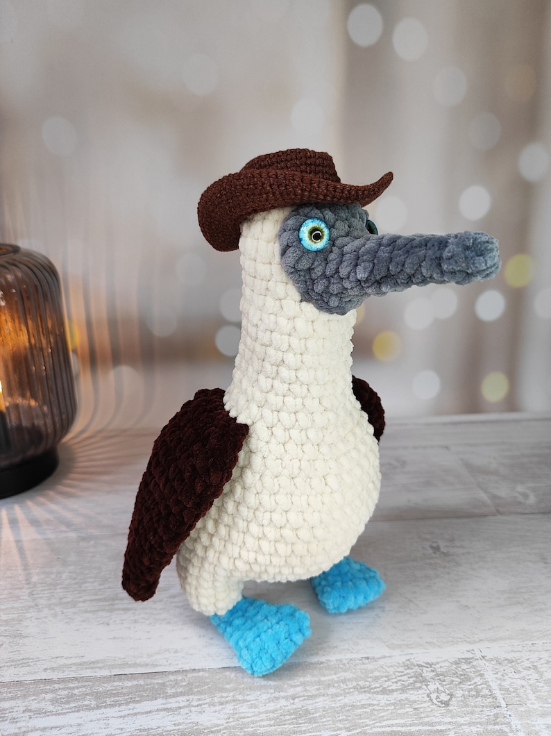 May include: A crocheted blue-footed booby wearing a brown cowboy hat. The bird is white with brown wings and blue feet.