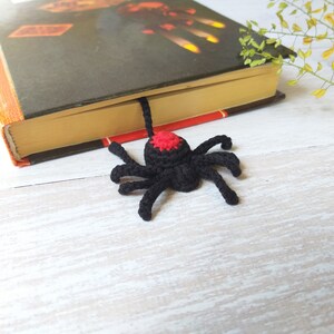 K&ouml;nnte beinhalten: Ein schwarzer, geh&auml;kelter Spinnen-Lesezeichen mit rotem Akzent auf dem Kopf liegt auf einem Buch. Das Lesezeichen ist handgefertigt und verleiht jedem Leseerlebnis eine einzigartige Note. Das Buch hat einen orangefarbenen und schwarzen Einband.