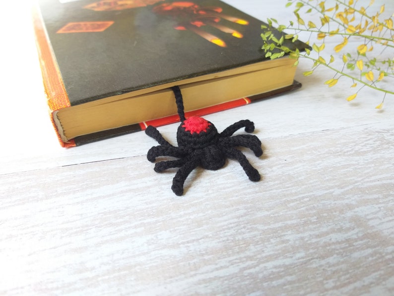 K&ouml;nnte beinhalten: Ein schwarzer, geh&auml;kelter Spinnen-Lesezeichen mit rotem Akzent auf dem Kopf liegt auf einem Buch. Das Lesezeichen ist handgefertigt und verleiht jedem Leseerlebnis eine einzigartige Note. Das Buch hat einen orangefarbenen und schwarzen Einband.