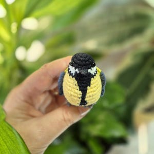 May include: A handmade crochet bird with a yellow body, black head, and blue wings. The bird has white accents on its face and a black stripe down its chest. The bird is a small, decorative item.