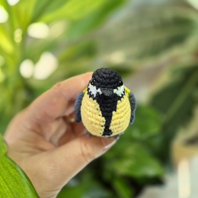 May include: A handmade crochet bird with a yellow body, black head, and blue wings. The bird has white accents on its face and a black stripe down its chest. The bird is a small, decorative item.