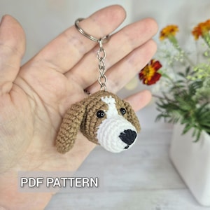 Può includere: Un portachiavi a forma di testa di cane all'uncinetto, marrone, bianco e nero. La testa del cane è dettagliata con occhi neri e una macchia bianca sulla fronte e sul muso. Il portachiavi include un anello e una catena di metallo. Il testo sull'immagine recita "PDF PATTERN".