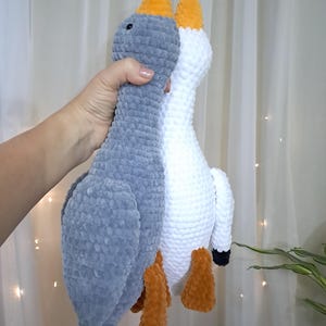 May include: Two crocheted goose plushies, one gray and one white, with orange beaks and legs. The plushies are standing upright and are facing each other.
