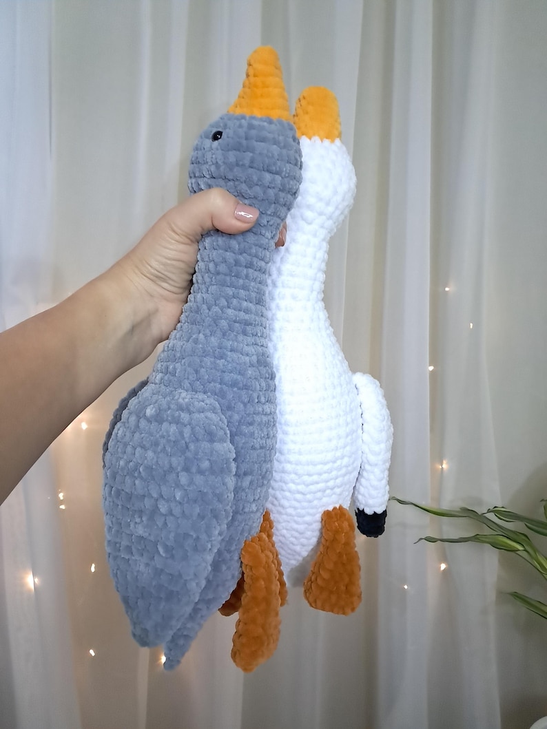 May include: Two crocheted goose plushies, one gray and one white, with orange beaks and legs. The plushies are standing upright and are facing each other.