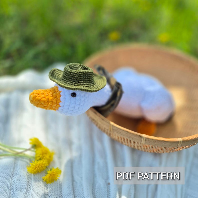 May include: A white crocheted goose wearing a green crocheted hat, sitting in a woven basket. The goose has a yellow beak and orange feet. The basket is on a white and yellow blanket. The text "PDF PATTERN" is visible in the bottom right corner of the image.