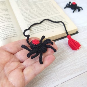 K&ouml;nnte beinhalten: Schwarzer und roter H&auml;kel-Spinnen-Lesezeichen mit roter Quaste. Die Spinne hat einen roten Fleck auf dem R&uuml;cken und wird in einer Hand in der N&auml;he eines offenen Buches gehalten. Zus&auml;tzliche Spinnen befinden sich im Hintergrund.