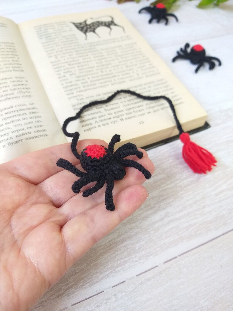 K&ouml;nnte beinhalten: Schwarzer und roter H&auml;kel-Spinnen-Lesezeichen mit roter Quaste. Die Spinne hat einen roten Fleck auf dem R&uuml;cken und wird in einer Hand in der N&auml;he eines offenen Buches gehalten. Zus&auml;tzliche Spinnen befinden sich im Hintergrund.
