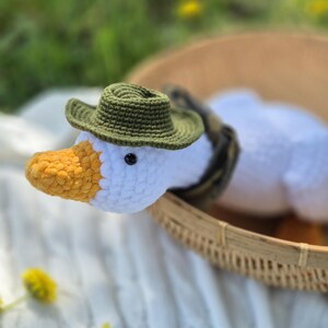 May include: A white crocheted goose wearing a green crocheted hat. The goose is sitting in a woven basket on a white crocheted blanket.