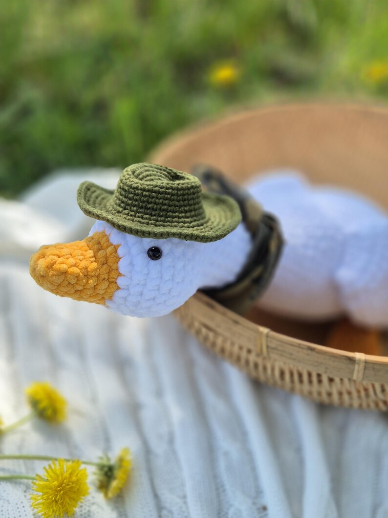 May include: A white crocheted goose wearing a green crocheted hat. The goose is sitting in a woven basket on a white crocheted blanket.