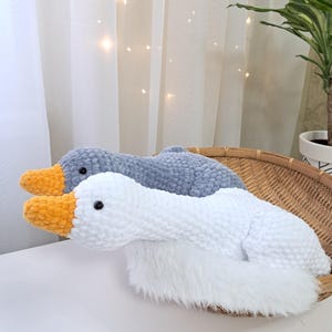 May include: Two plush goose-shaped pillows. One is white with an orange beak, the other is gray with an orange beak. Both have black eyes and are made of soft, textured material. They rest in a woven basket.