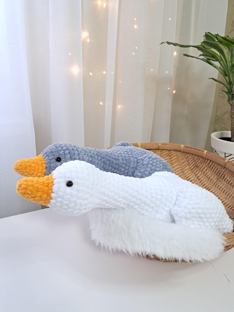 May include: Two plush goose-shaped pillows. One is white with an orange beak, the other is gray with an orange beak. Both have black eyes and are made of soft, textured material. They rest in a woven basket.
