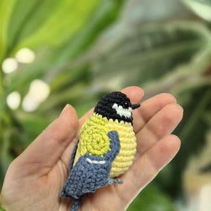 May include: A handmade crochet bird figurine. The bird has a yellow body, a black head, and blue wings. The bird is held in a hand against a blurred green background. The bird is a miniature, detailed craft item.
