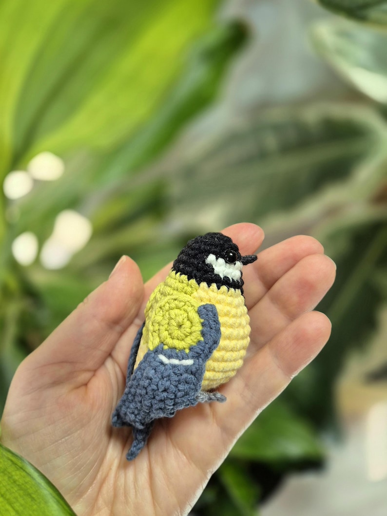 May include: A handmade crochet bird figurine. The bird has a yellow body, a black head, and blue wings. The bird is held in a hand against a blurred green background. The bird is a miniature, detailed craft item.