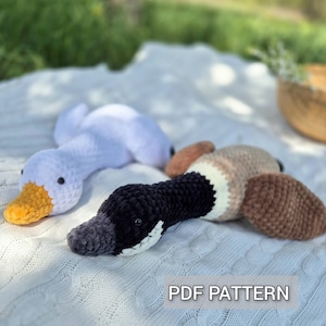 Può includere: Due peluche di anatra all'uncinetto, uno bianco con becco giallo, l'altro nero e marrone. Il testo "PDF PATTERN" è visibile. Questi giocattoli fatti a mano sono perfetti per una cameretta o come regalo unico.