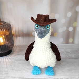 May include: A crocheted blue-footed booby wearing a brown cowboy hat. The bird is white with brown accents and blue feet.