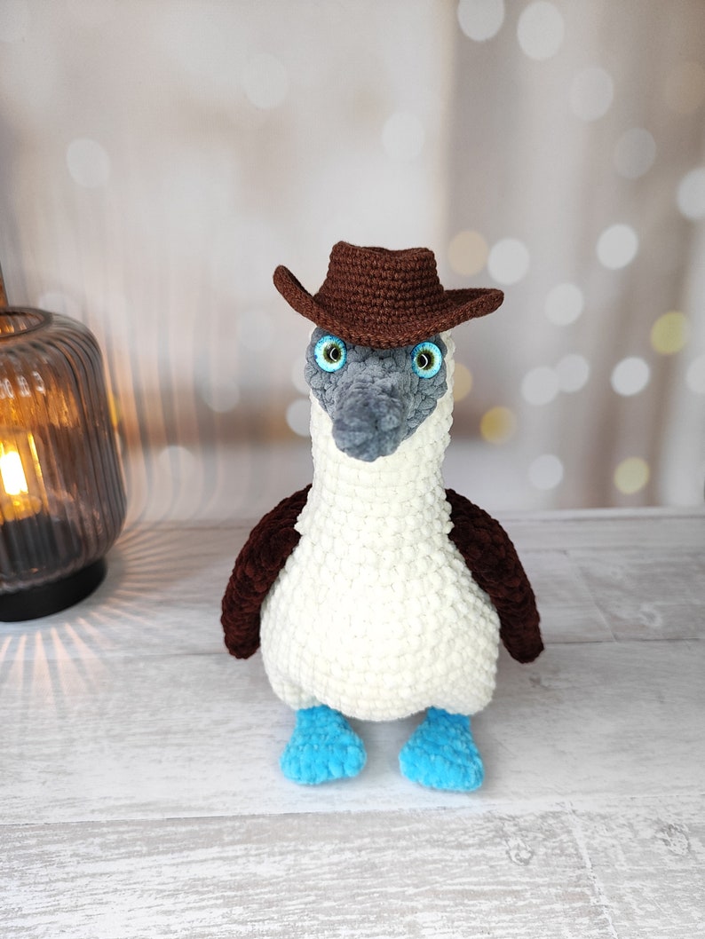 May include: A crocheted blue-footed booby wearing a brown cowboy hat. The bird is white with brown accents and blue feet.