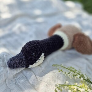 May include: A handmade crochet duck toy. The duck features a dark blue head and neck, a white and cream-colored patch, and brown wings. The toy is lying on a white, textured blanket.
