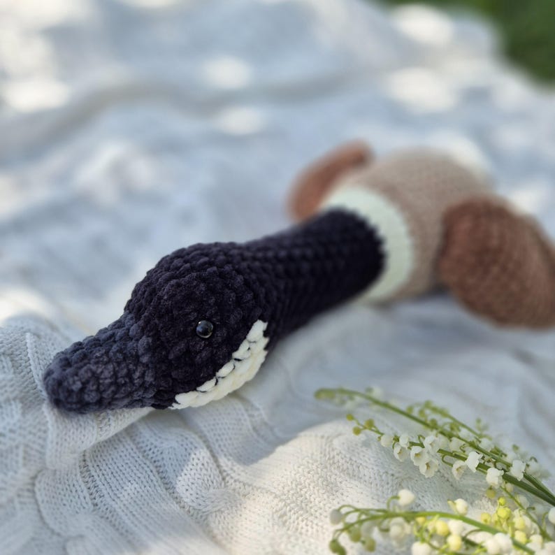 May include: A handmade crochet duck toy. The duck features a dark blue head and neck, a white and cream-colored patch, and brown wings. The toy is lying on a white, textured blanket.