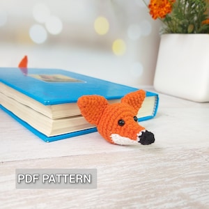 May include: A crocheted fox bookmark with orange fur, white accents, and black eyes. The bookmark is placed on a blue book with the text "PDF PATTERN" visible below.