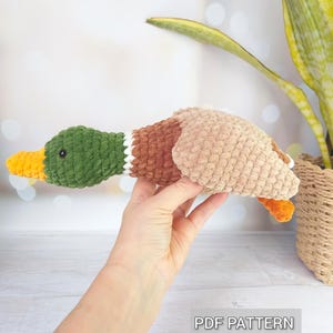May include: A crocheted mallard duck plush toy featuring a dark green head, a bright orange beak, a white neck ring, a brown chest, and a light tan body with orange feet. The item is labeled as a PDF PATTERN in the bottom right corner.