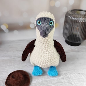 May include: A crocheted blue-footed booby bird plush toy with brown wings, white body, and blue feet. The toy is sitting on a white wooden surface with a brown crocheted hat beside it.