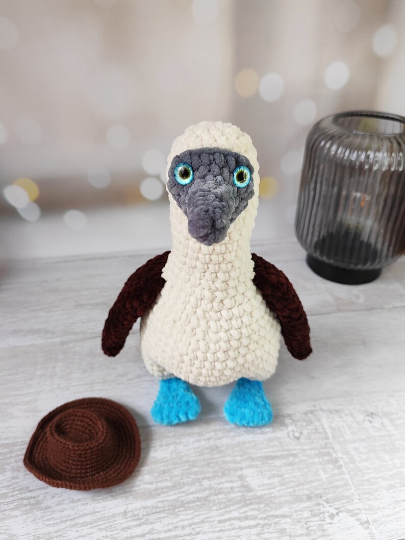May include: A crocheted blue-footed booby bird plush toy with brown wings, white body, and blue feet. The toy is sitting on a white wooden surface with a brown crocheted hat beside it.
