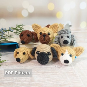 Op de afbeelding: Zes gehaakte hondenbladwijzers in verschillende rassen, waaronder een mops, een corgi, een poedel en een teckel. De bladwijzers liggen op een boek met een blauwe rug.