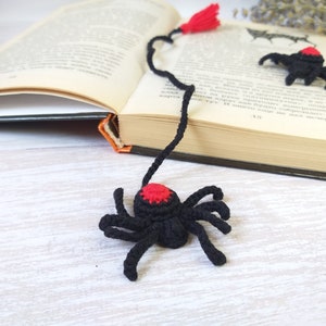 K&ouml;nnte beinhalten: H&auml;kel-Lesezeichen in Form einer schwarzen Spinne mit rotem Akzent und rotem Quastenanh&auml;nger. Das Lesezeichen liegt auf einem offenen Buch, im Hintergrund eine helle Holzoberfl&auml;che. Eine weitere Spinne ist ebenfalls zu sehen.