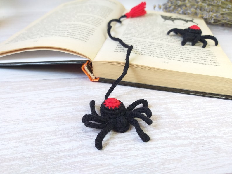 K&ouml;nnte beinhalten: H&auml;kel-Lesezeichen in Form einer schwarzen Spinne mit rotem Akzent und rotem Quastenanh&auml;nger. Das Lesezeichen liegt auf einem offenen Buch, im Hintergrund eine helle Holzoberfl&auml;che. Eine weitere Spinne ist ebenfalls zu sehen.