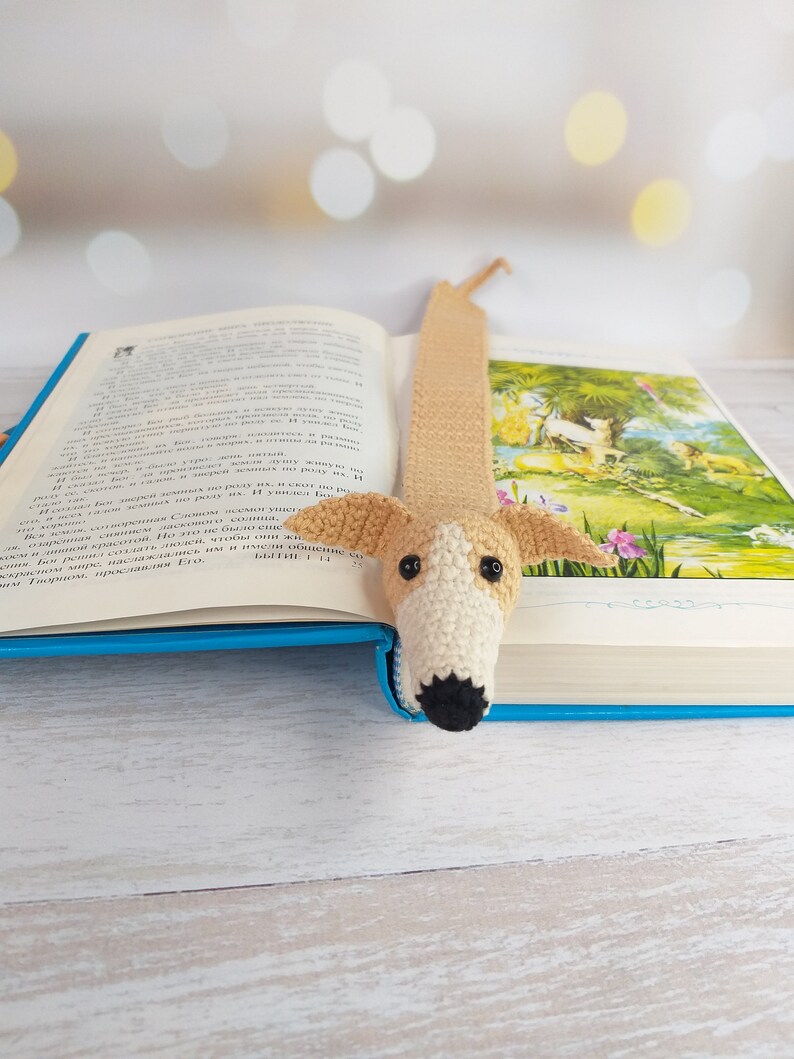 Może przedstawiać: Zakładka do książki w kształcie psa, wykonana na szydełku, w kolorze beżowym i białym z czarnymi oczami i nosem. Zakładka znajduje się w otwartej książce z niebieską okładką.
