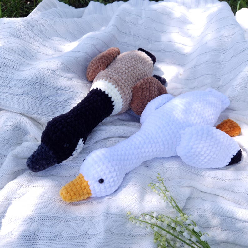 May include: Two plush, handmade bird toys. One is a white goose with a yellow beak and black accents. The other is a duck with a black head, brown wings, and a tan body. Both are lying on a white, textured blanket.