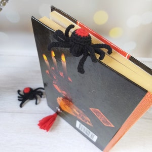 K&ouml;nnte beinhalten: Ein schwarzer H&auml;kel-Spinnen-Lesezeichen mit rotem Akzent liegt auf einem Buch. Das Buch hat einen schwarzen Einband mit orangefarbenen und roten Details. Zwei weitere Spinnen-Lesezeichen und eine rote Quaste sind ebenfalls zu sehen.