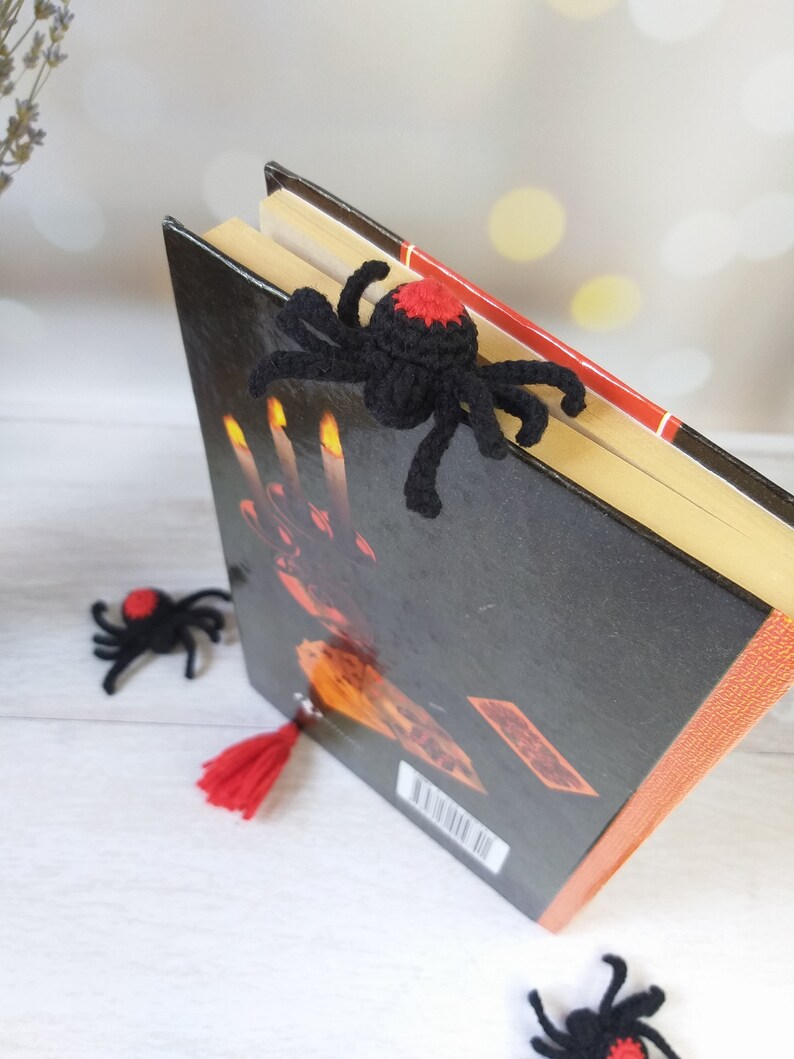 K&ouml;nnte beinhalten: Ein schwarzer H&auml;kel-Spinnen-Lesezeichen mit rotem Akzent liegt auf einem Buch. Das Buch hat einen schwarzen Einband mit orangefarbenen und roten Details. Zwei weitere Spinnen-Lesezeichen und eine rote Quaste sind ebenfalls zu sehen.