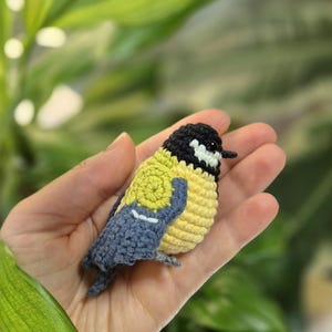 May include: A handmade crochet bird in yellow, black, and blue. The small bird has a yellow body, a black head, and blue wings. The bird is held in a hand against a blurred green background. The bird is a decorative item.