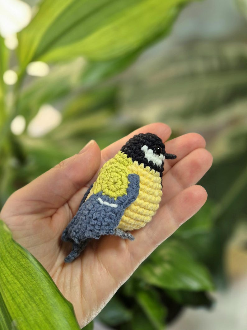 May include: A handmade crochet bird in yellow, black, and blue. The small bird has a yellow body, a black head, and blue wings. The bird is held in a hand against a blurred green background. The bird is a decorative item.