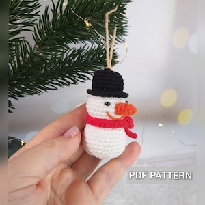 以下が含まれることがあります： 黒い帽子、赤いマフラー、オレンジ色の鼻をした、かぎ針編みで作られた雪だるまのオーナメント。オーナメントは木の枝にぶら下がっています。