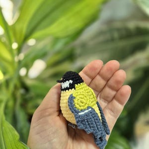 May include: A handmade crochet bird in yellow, black, and blue. The small bird has a detailed design, with a black head, yellow body, and blue wings. The bird is held in a person's hand, with a blurred green background.