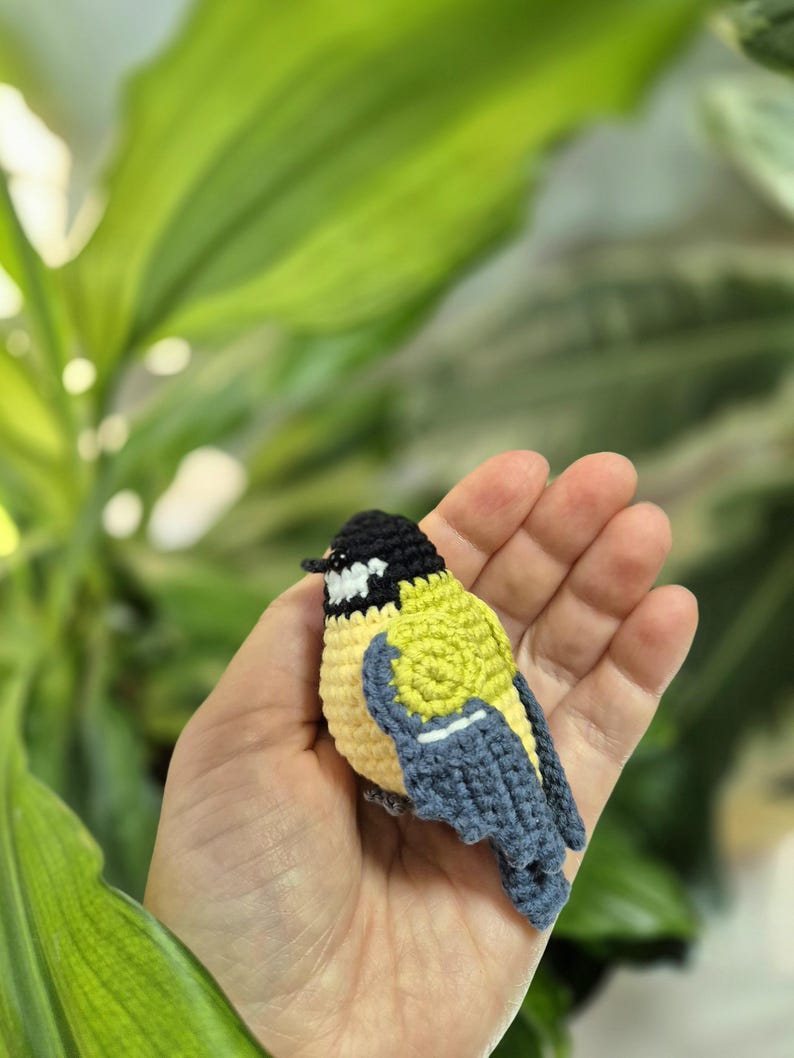 May include: A handmade crochet bird in yellow, black, and blue. The small bird has a detailed design, with a black head, yellow body, and blue wings. The bird is held in a person's hand, with a blurred green background.