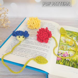 May include: Crocheted flower bookmarks in yellow, red, and blue, with green leaf accents, rest on an open book. The bookmarks are handmade and feature a detailed floral design. The text "PDF PATTERN" is visible.