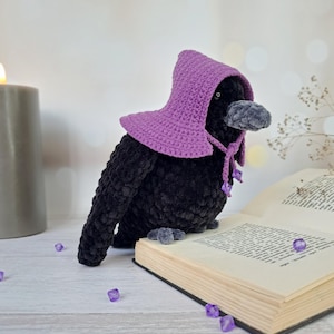 May include: A black plush crow toy with a textured, velvety finish, wearing a purple crocheted hood that ties under the beak with small purple beads. The crow is positioned on an open book, featuring a dark grey beak and small dark eyes.
