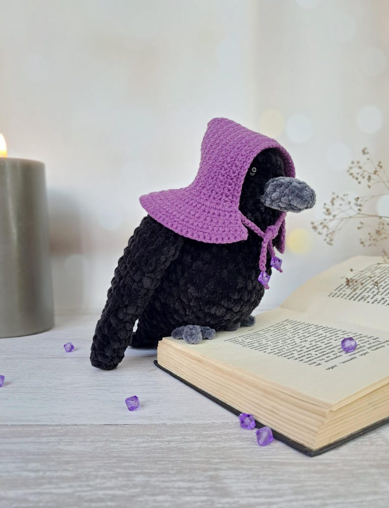 May include: A black plush crow toy with a textured, velvety finish, wearing a purple crocheted hood that ties under the beak with small purple beads. The crow is positioned on an open book, featuring a dark grey beak and small dark eyes.