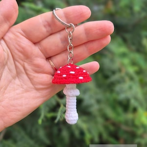 May include: A crocheted mushroom keychain featuring a bright red cap with white spots and a white stem. The mushroom is attached to a silver-colored metal split ring by a short chain. The text PDF PATTERN is visible in the bottom right corner.