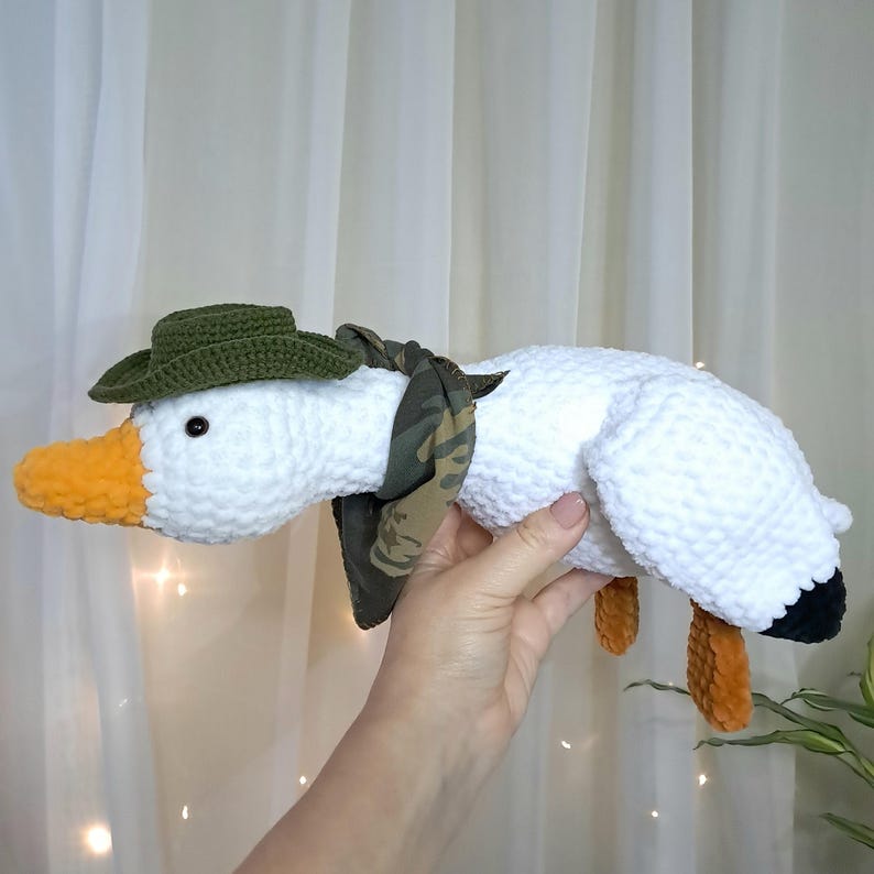 May include: White crocheted goose plush toy wearing a green hat and a camouflage scarf. The goose has orange feet and a black beak.